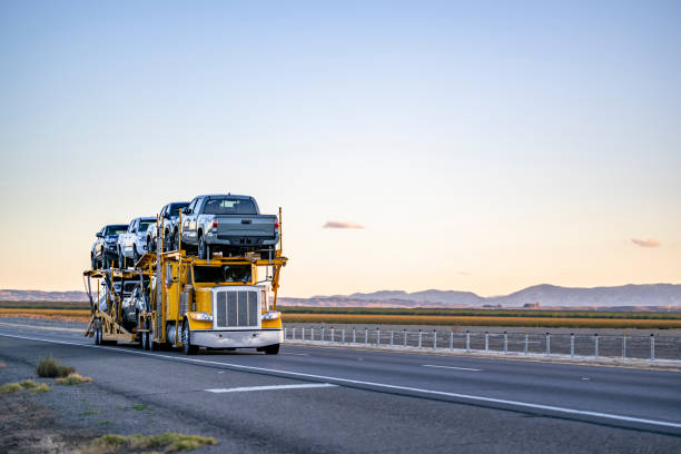 A yellow car carrier truck loaded with several vehicles drives on a highway with mountains and open fields in the background under a clear sky, exemplifying reliable NY Car Shipping Bay Shore services.