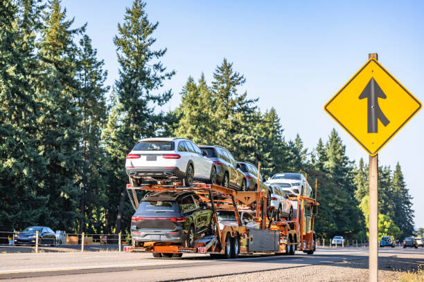 A NY Car Shipping Bay Shore carrier truck transporting multiple vehicles drives on a road surrounded by trees, with a yellow merge traffic sign visible on the right side.