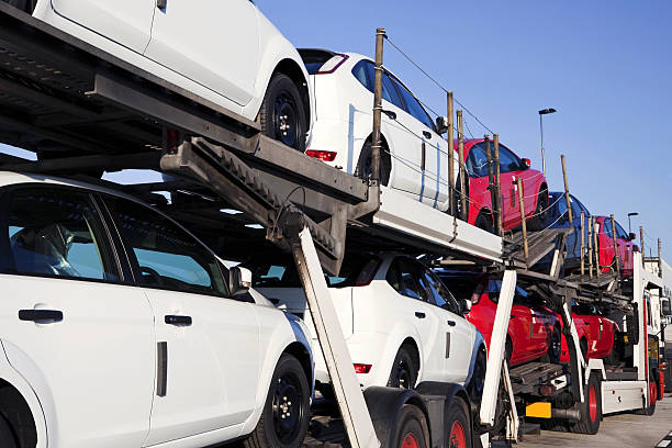 A car carrier truck, operated by NY Auto Transport Bay Shore, transports multiple white and red vehicles stacked on two levels against a clear blue sky in the background.